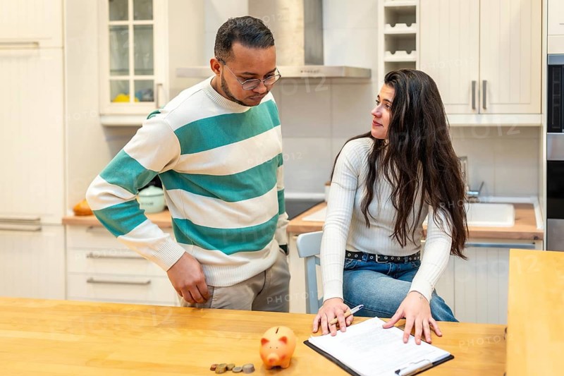 Young couple sitting and standing in their kitchen, seriously discussing household finances, managing monthly budget, and planning their financial future with a piggy bank and documents on the table