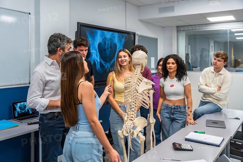 Anatomy teacher explaining the human skeleton to a diverse group of college students in a modern science classroom, utilizing an x ray display and interactive learning methods