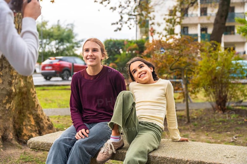 Two sisters smiling and relaxing on a park bench while a family member photographs their candid, joyful moment of togetherness and everyday outdoor bonding