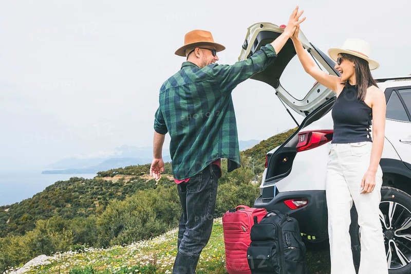 Happy Travelers tourist couple hi5 stand put luggage to white rented SUV vehicle trunk on scenic Mediterranean viewpoint. Weekend get away road trip arrival, travel preparation, planning holiday\