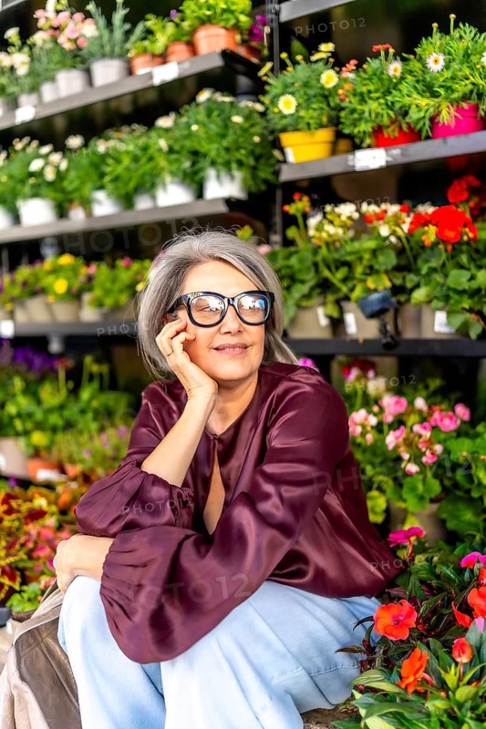 Mature woman with gray hair and glasses smiling thoughtfully, surrounded by colorful potted flowering plants and green foliage in a garden center environment