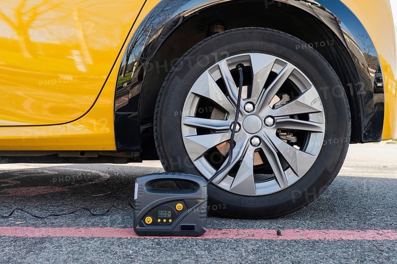 An electric car's wheel is being inflated using a portable tire inflator on an urban street. The close-up highlights the modern design of the alloy wheel and inflation device