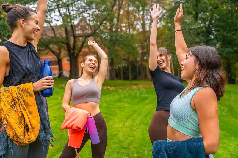 Group of enthusiastic young women friends raising their arms in celebration after a successful fitness workout or yoga session in a lush green park, sharing joy and friendship