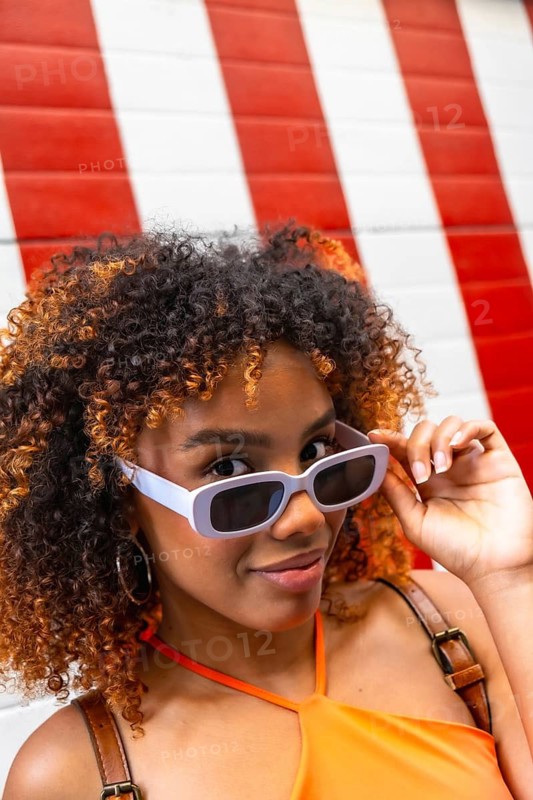 Young black woman with curly hair confidently adjusting her stylish sunglasses, looking directly at the viewer with a friendly expression against a vibrant red and white striped background