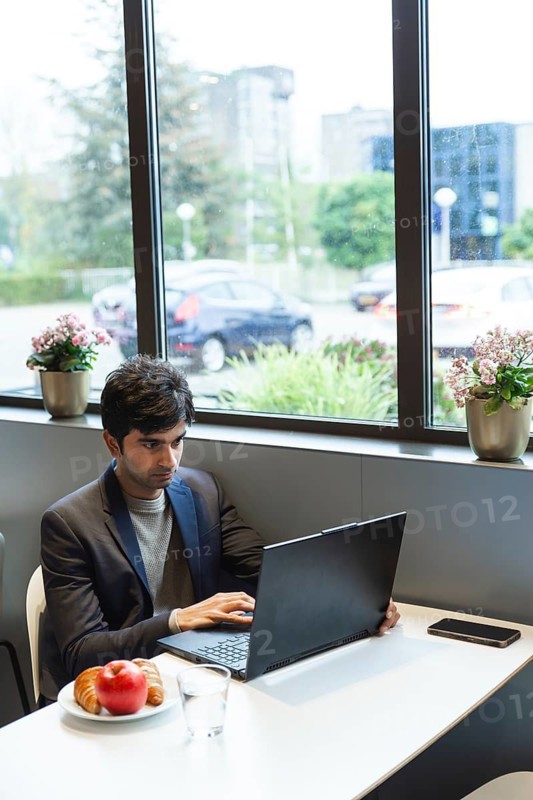 A indian man sits in a hotel cafe, focused on his laptop at a conference of AI in Amsterdam. Sunlight filters through the window, creating a serene atmosphere. A healthy breakfast sits nearby