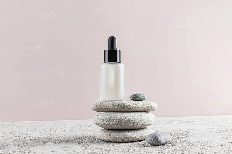 A frosted glass essential serum bottle with a dropper cap, balanced on smooth pebbles against a neutral background, evokes calmness and simplicity in skincare