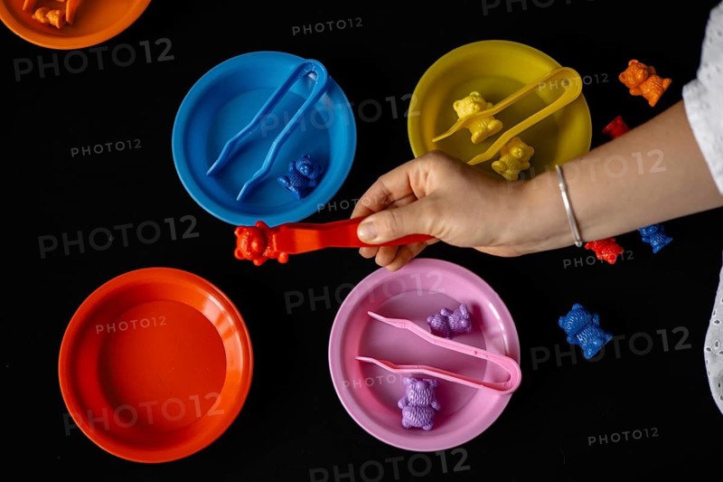 Toy bears are sorted into colorful plates with tongs by a hand on black background