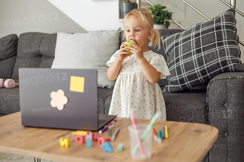 A young Caucasian girl with blonde hair stands by a laptop on a wooden table. Colorful toys and stationery are scattered around her in a cozy living room setting