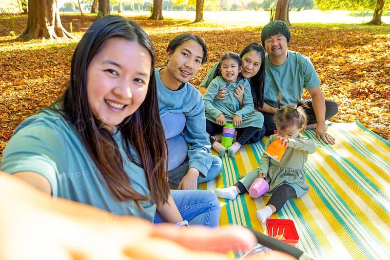 Happy Asian family having a joyful picnic outdoors, sitting on a blanket surrounded by autumn leaves. They're capturing the moment with a selfie, sharing smiles and laughter