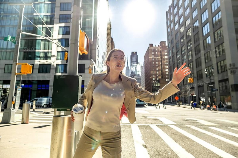Busy businesswoman with a travel mug actively hailing a taxi on a sunny day in manhattan, navigating the dynamic urban environment with determination and purposeful action