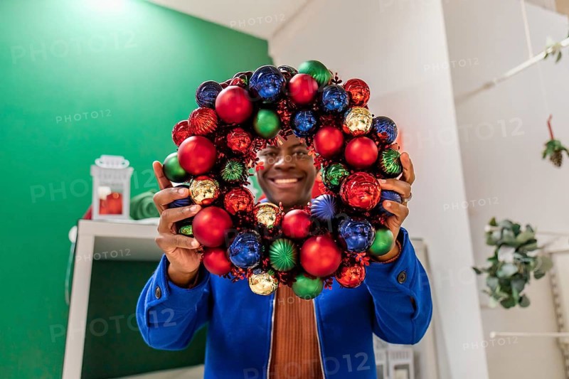 Close up young happy fun african man 20s in sweater look through Advent wreath on green background. Happy New Year celebration concept.