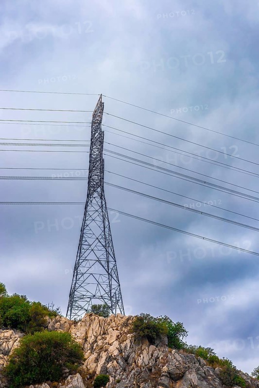 A tall electricity transmission tower stands on a rocky hill. Power lines stretch across the sky under a cloudy atmosphere. The scene emphasizes energy infrastructure