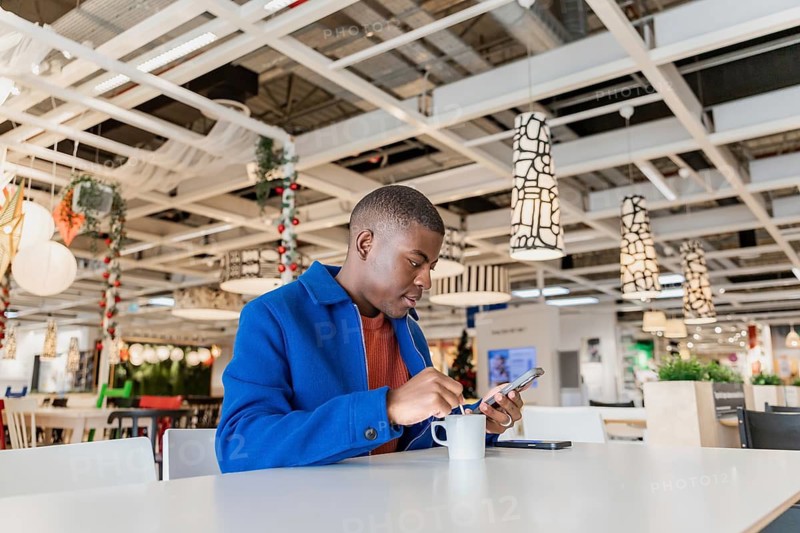 A young Black man using a phone and holding a cup and sits at a table in a modern cafe, The interior features contemporary decor and bright lighting