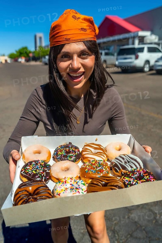 Woman standing outdoors on a sunny day, holding and offering a box filled with a variety of colorful glazed and sprinkled donuts, expressing joy and sweetness