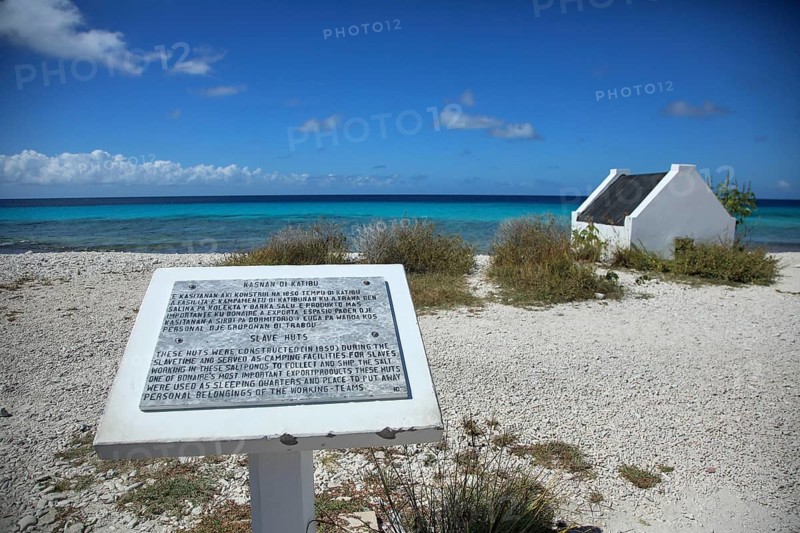 Information board on the pebble beach with a small white house and turquoise sea in the background under a blue sky, historic white slave huts on the coast of Bonaire, the Netherlands Antilles and the Caribbean
