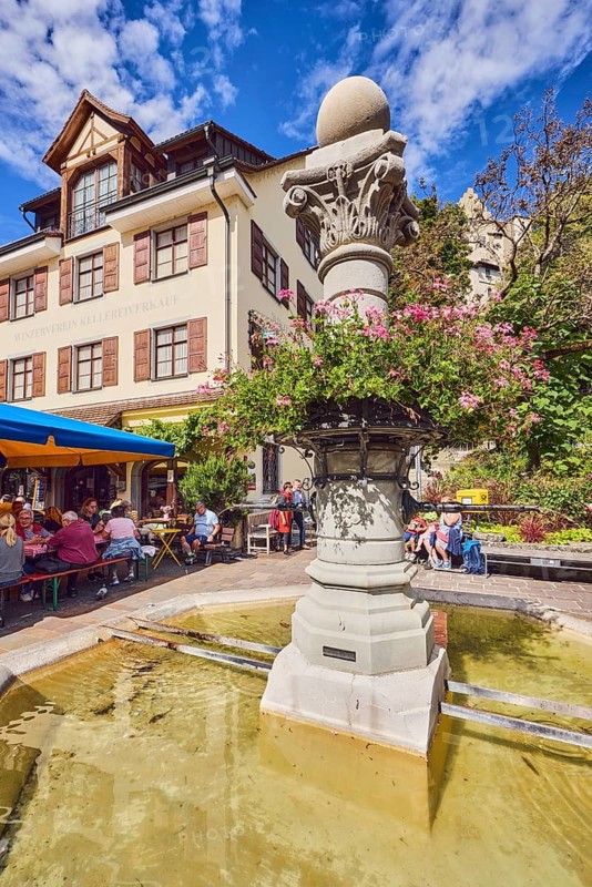 Ancient fountain, historic city fountain, flower decoration, general architecture, houses, public benches, outdoor area of a restaurant, wine shop, shopping, winegrowers' association winery sales, pedestrians, seated people and gastronomic visitors as a secondary motif, trees, blue sky, altocumulus clouds, Unterstadtstraße, Meersburg, Lake Constance district, Baden-Württemberg, Germany