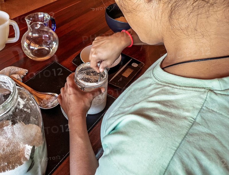 A woman in a light shirt mixes a sourdough starter in a jar for make bread on a wooden kitchen counter, surrounded by various tools and ingredients used for baking