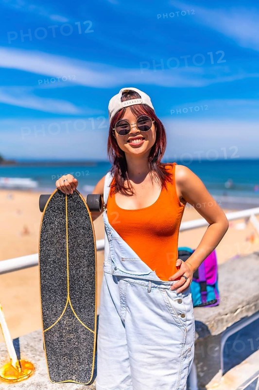 Vertical Portrait of a cool young female asian skater in summer modern clothes, cap and sunglasses smiling at camera standing next to the beach