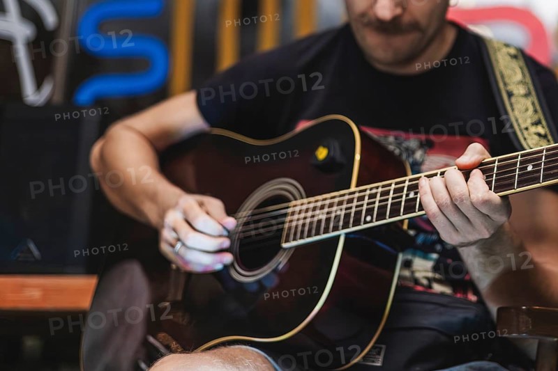 A young Caucasian man with a beard plays an acoustic guitar. He wears a black t-shirt and is focused on his music in a casual setting