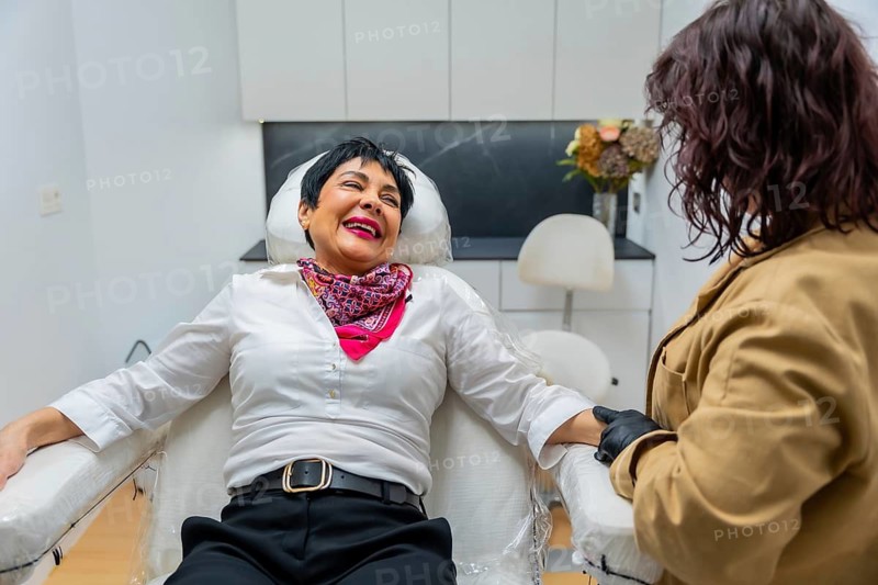 Mature woman smiling broadly while an aesthetician, wearing protective gloves, prepares her arm for a cosmetic procedure in a modern medical spa specializing in skin care and anti aging treatments
