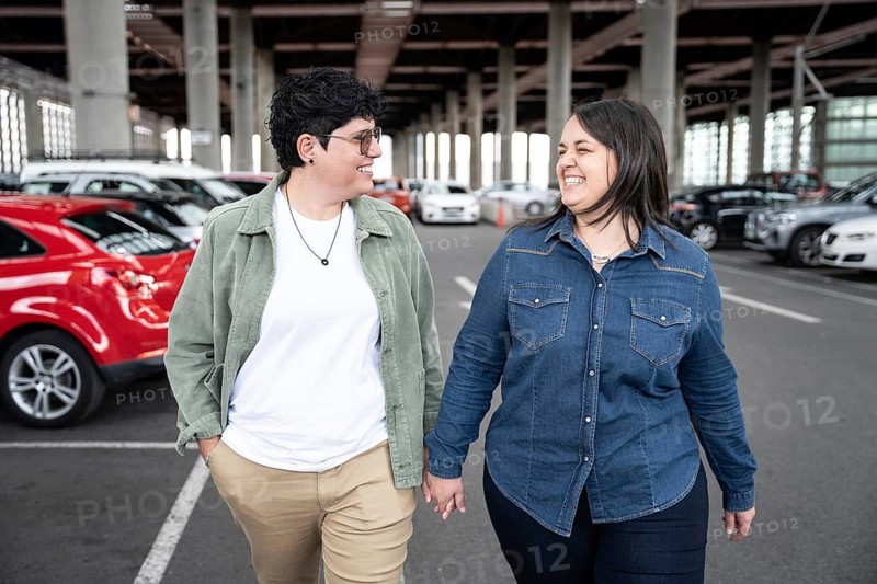 Lesbian couple laughing and walking hand in hand through a parking area. They are dressed casually, exuding happiness and connection in an urban setting