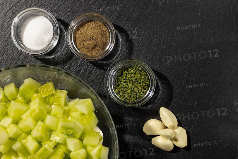 Bowl with sliced cucumber and sour cream, spices and garlic next to it