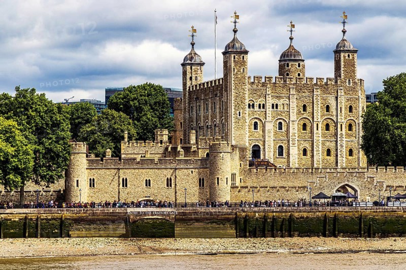 Visitors line the riverside path beside the Tower of London, where the pale turrets of the Norman White Tower rise behind concentric medieval walls that once served the fortress as royal palace, treasury, mint and prison across nine centuries, London, England, United Kingdom