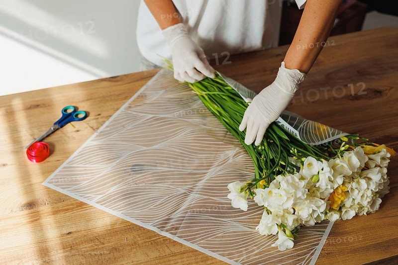 A person in gloves carefully arranges a bunch of fresh flowers, combining vibrant yellow and white blooms. The setting is a bright, airy workspace, enhancing the natural beauty of the flowers