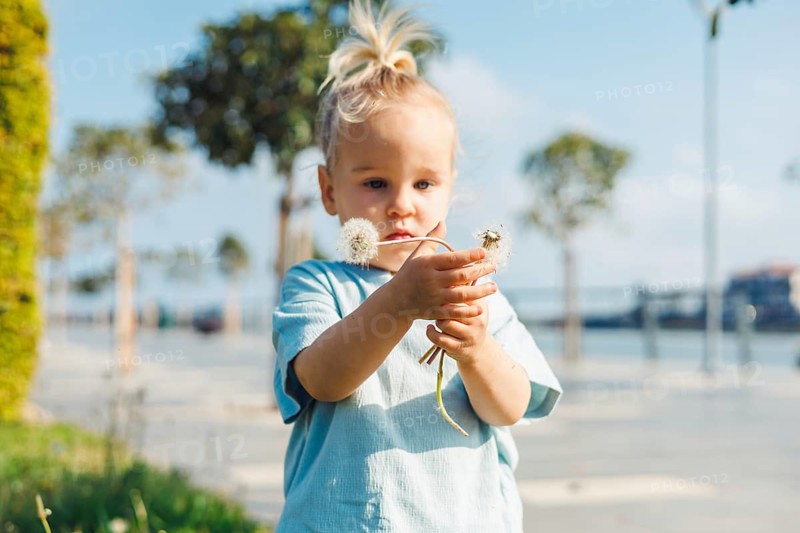 Young child toddler girl stands in a vibrant park on a sunny day, exploring nature with curious eyes. Kid holds a delicate flower, surrounded by greenery and a peaceful waterfront view