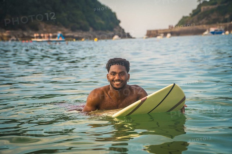 Young black man in the ocean smiling at the camera, holding a surfboard on a sunny summer day, reflecting a carefree lifestyle and water sports activity