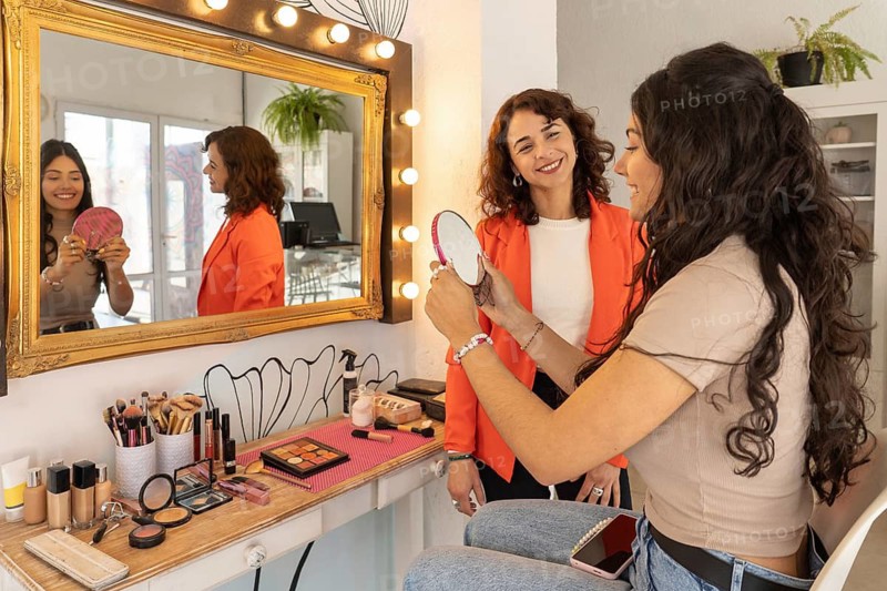 Satisfied client smiles while checking her completed makeup look in a hand mirror as the makeup artist stands beside her in a beauty salon