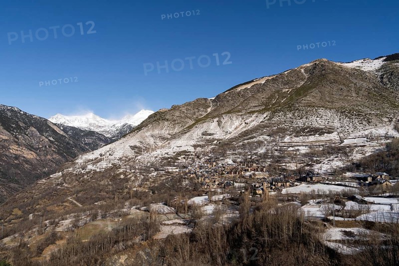 A quaint mountain village sits surrounded by steep, snowy peaks under a clear blue sky, embodying the serene, unspoiled beauty of rural life