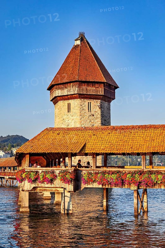 Medieval wooden bridge with tower and blooming flowers over a body of water in sunny weather, The Chapel Bridge in Lucerne in Switzerland