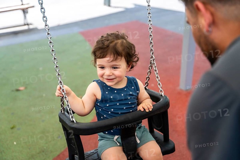 Happy infant child sitting in a safety swing, looking at parent and smiling widely during playtime, enjoying an outdoor family moment in a public park