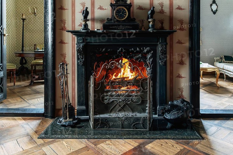 A vintage room features an ornate dark fireplace with a bright burning fire and a wrought iron guard. A black clock and two sculpted birds sit on the mantel. Fireplace tools stand to the side. Red patterned wallpaper and wooden parquet flooring add warmth. Through the doorways, adjoining rooms with furniture are visible, creating a cozy and elegant interior