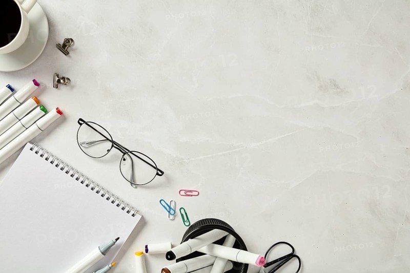 From above a neatly organized creative workspace featuring a sketchbook, colorful markers, glasses, and office supplies on a marble table, with a background of elegant white marble offering a clean aesthetic