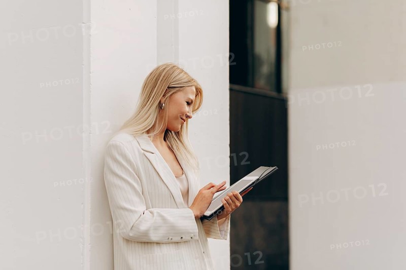 A businesswoman in a white blazer stands outside against a wall, looking at a digital tablet. She appears focused and engaged, capturing a moment of modern technology use