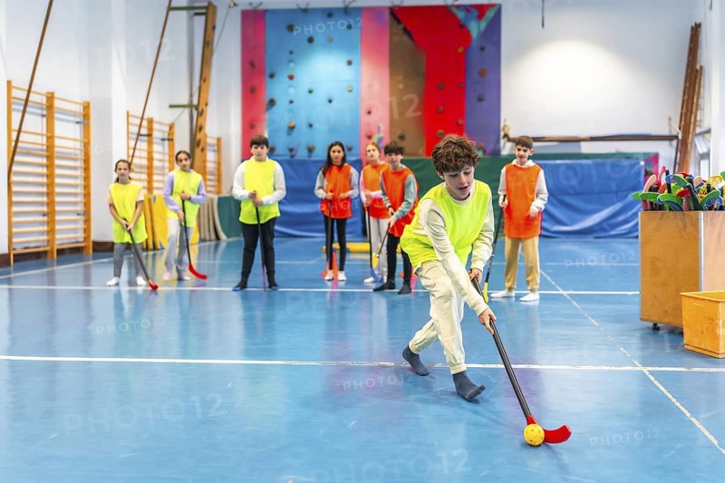 Group of elementary school students wearing bibs playing floorball in school gym during their physical education class