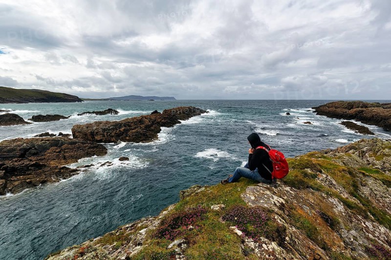 Hiker sitting on windy rocky coast, rain clouds, Melmore, Downings, Boyeeghter Strand, Murder Hole Beach, Sheephaven Bay, Rosguill Peninsula, Donegal, Ireland