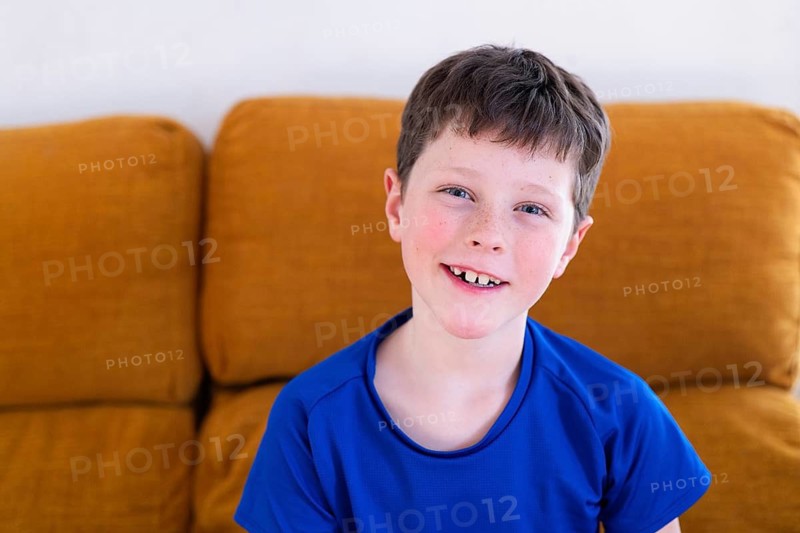 A young boy in a blue shirt, grinning with pride at home on a brown sofa, showing off his first missing milk tooth