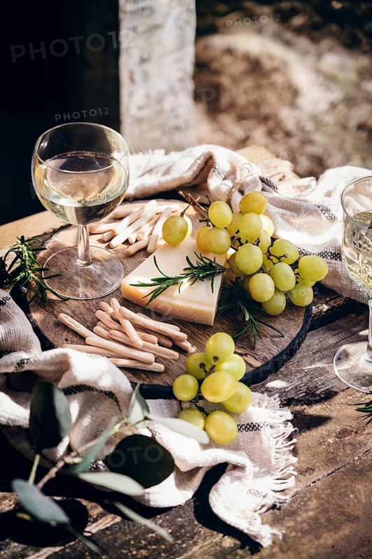 A elegant outdoor still life featuring a glass of white wine, green grapes, rosemary and cheese on a rustic wooden table. Sunlight highlights the texture and arrangement, creating a inviting ambiance