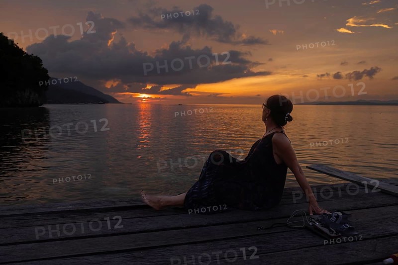 A mature female tourist relaxes on a wooden pier on Kri Island in the Raja Ampat Archipelago, Indonesia, admiring a breathtaking sunset over the calm sea