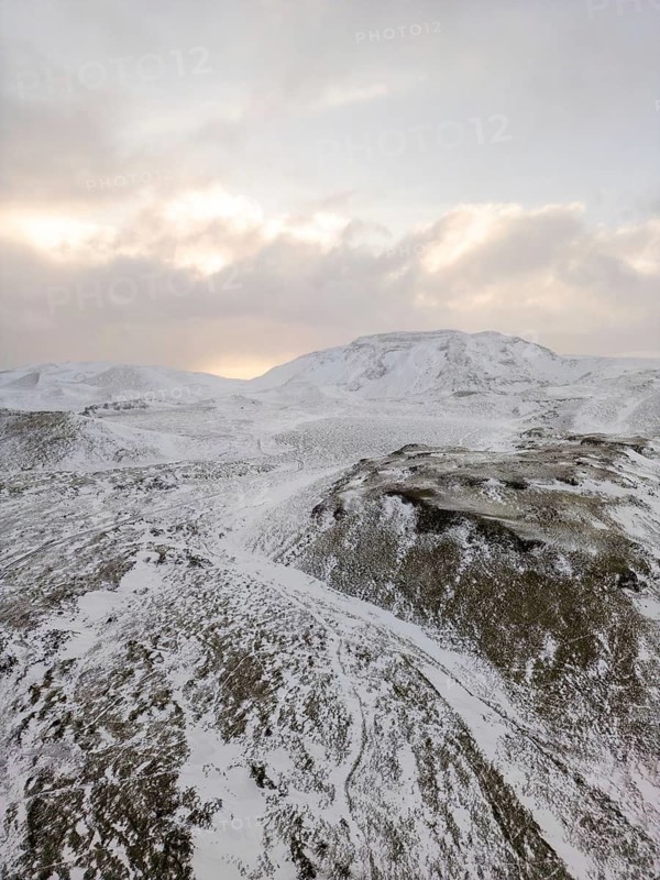 Aerial view of Iceland's winter landscape, showcasing snow-covered hills and distant mountains under a softly lit sunrise, highlighting the remote and wild nature of the terrain