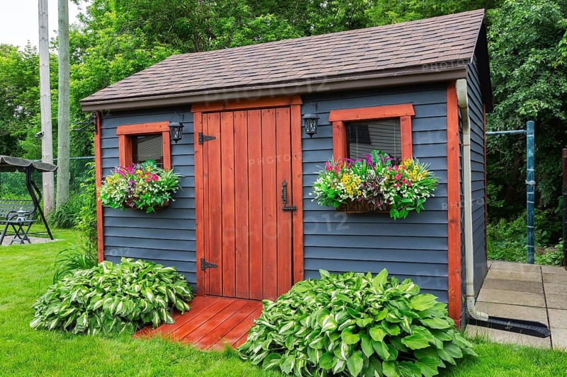 Dark grey clapboard cladded garden storage shed with cedar wood plank door and window flower boxes decorated with plastic flowers in summer, Quebec, Canada