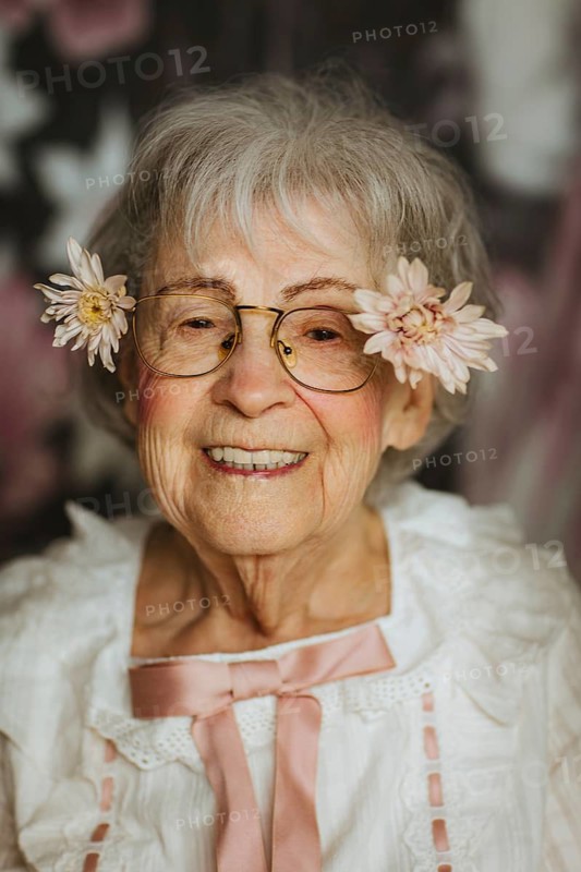 An elderly woman wearing glasses and flowers in her hair smiles warmly at the camera, showcasing joy and elegance in a studio setting with a floral backdrop