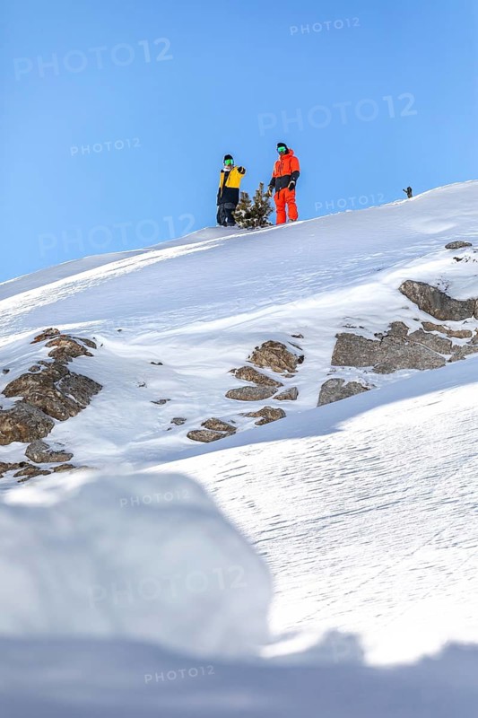 Two snowboarder friends in vibrant gear stand on a snowy mountain peak, poised to descend. Bright blue sky enhances the winter adventure setting, capturing the thrill of snowboarding