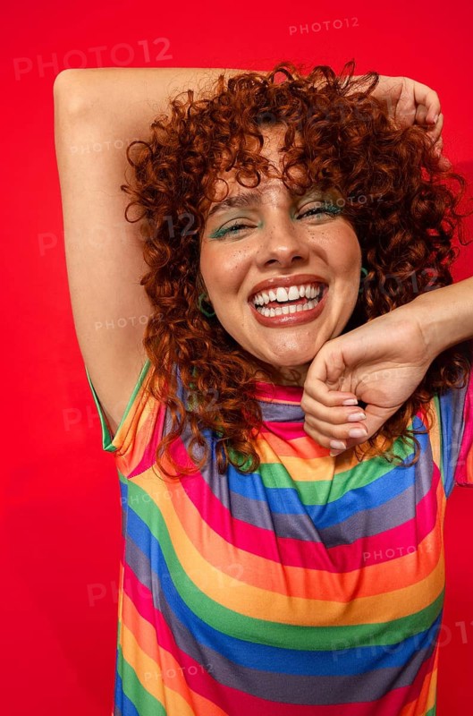 A cheerful woman with vibrant curly hair smiles broadly against a bold red background. She wears a rainbow-striped shirt, exuding joy and positivity in a lively pose