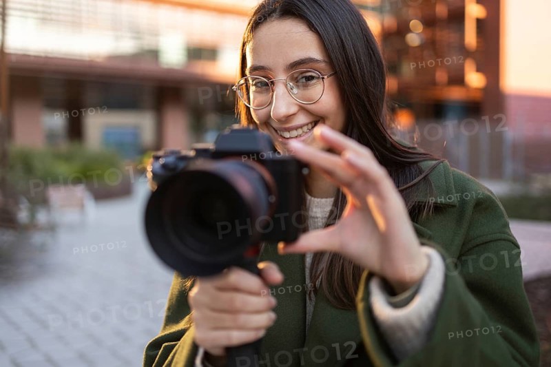 A smiling woman in a green coat holds a camera, capturing a moment outdoors at sunset She wears glasses and appears joyful, with a blurred modern building in the background