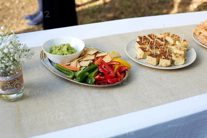 A beautifully decorated wedding table featuring a delicate floral arrangement in a glass jar, alongside platters of colorful vegetables, guacamole, and savory bites on a linen tablecloth