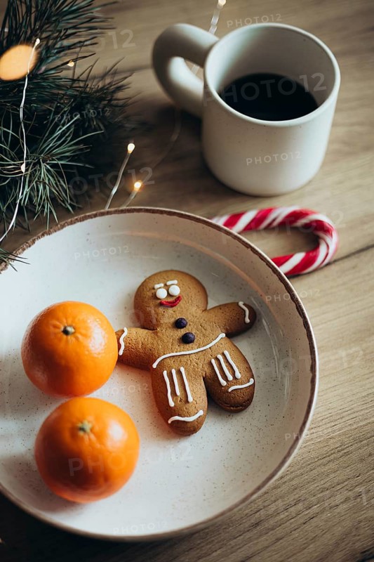 A joyful gingerbread man cookie on a plate with two oranges, accompanied by a candy cane, coffee, and festive lights Perfect for celebrating holiday cheer